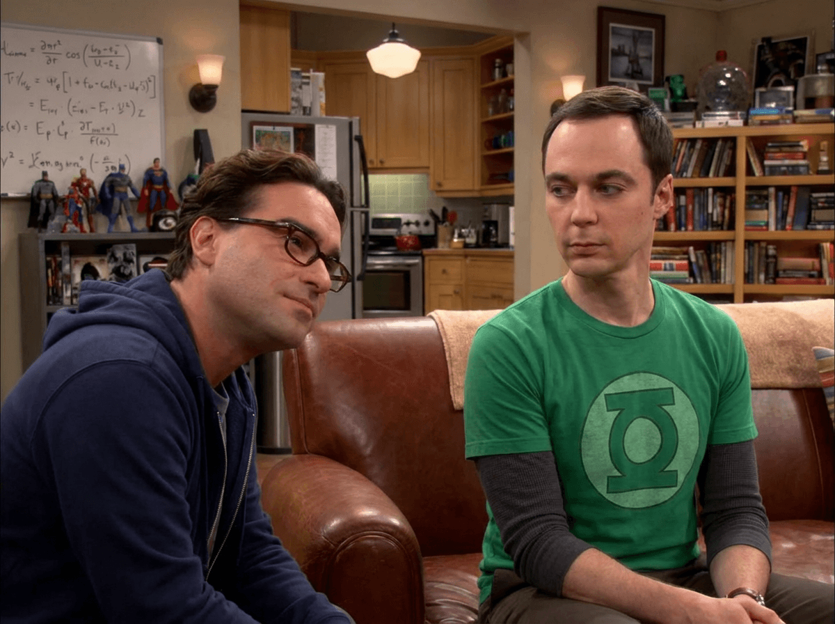 Leonard in a dark hoodie sits beside Sheldon in a green T-shirt on their brown sofa, with books and a whiteboard behind them.