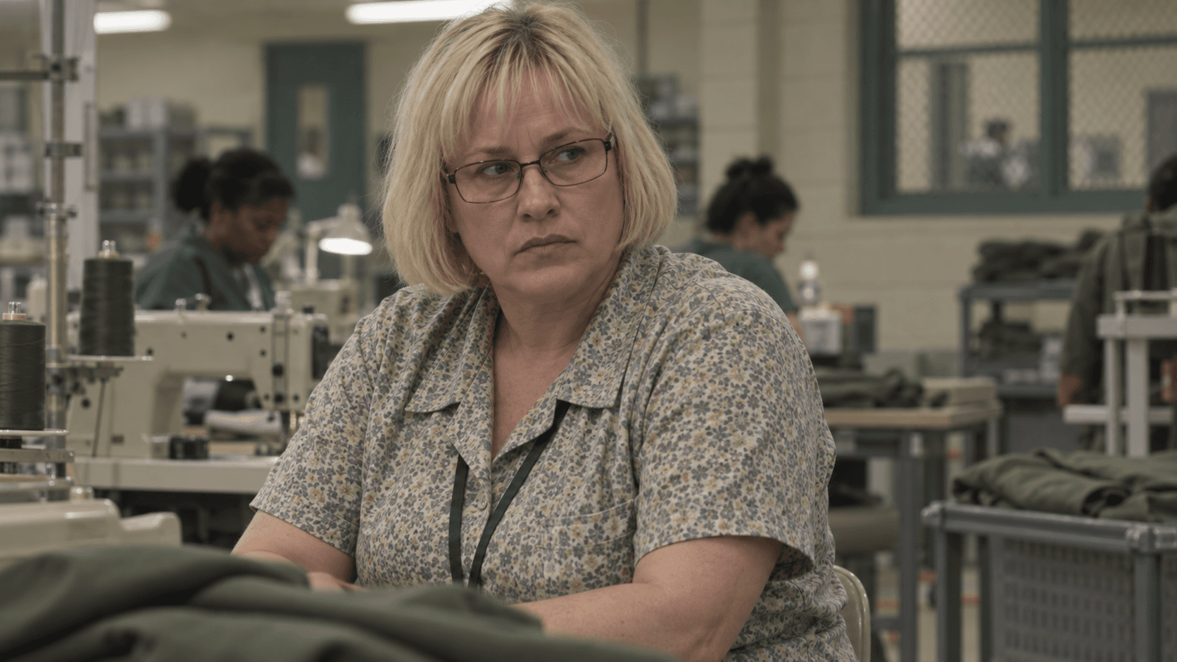 Patricia Arquette as Tilly Mitchell, with a blonde bob, glasses and patterned blouse, in a drab sewing workshop.