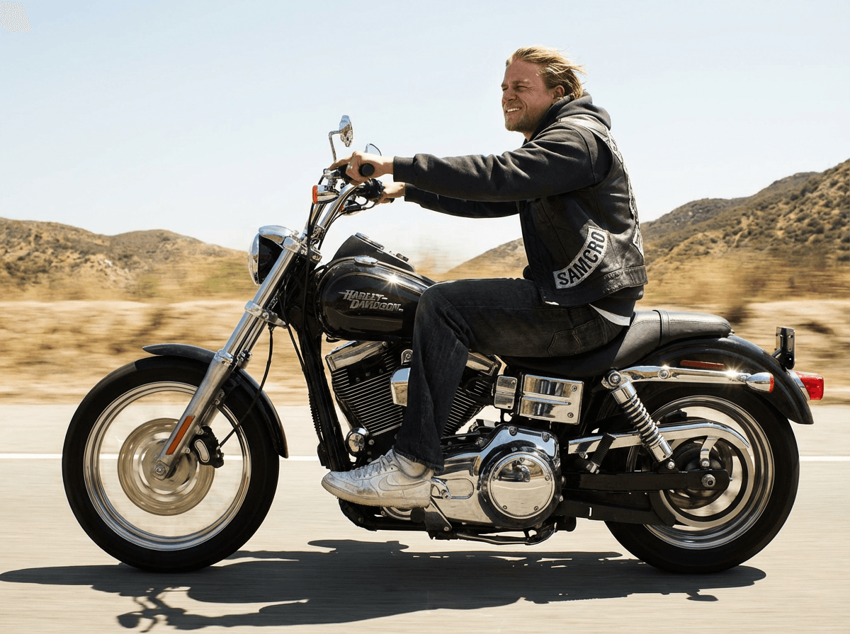 Jax Teller riding a black motorcycle past dry California hills in daylight.